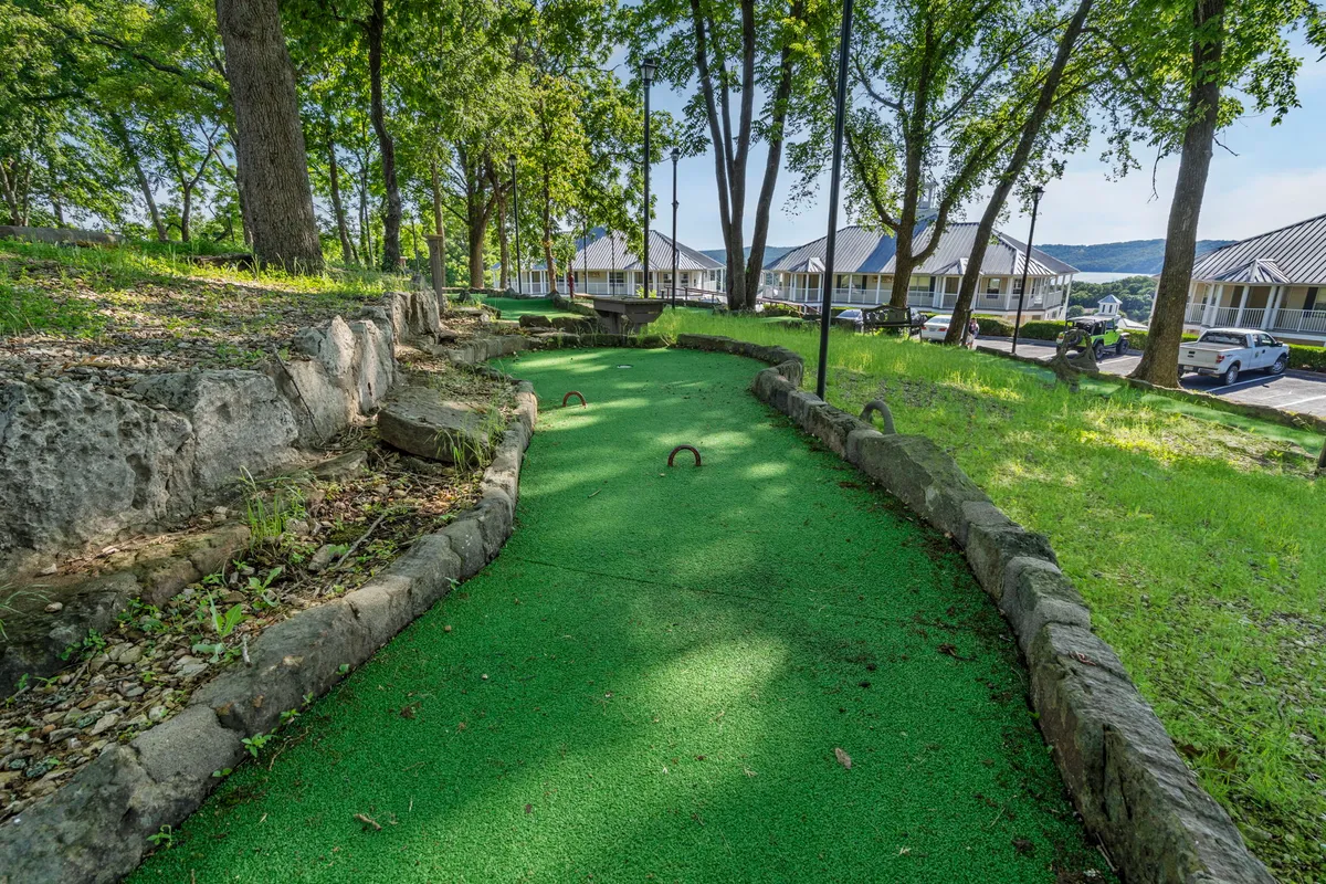 This image features a vibrant mini-golf course with green turf, surrounded by mature trees and natural scenery. The course looks engaging and is clearly designed for friendly, family-oriented play, inviting groups to gather for some lighthearted competition.