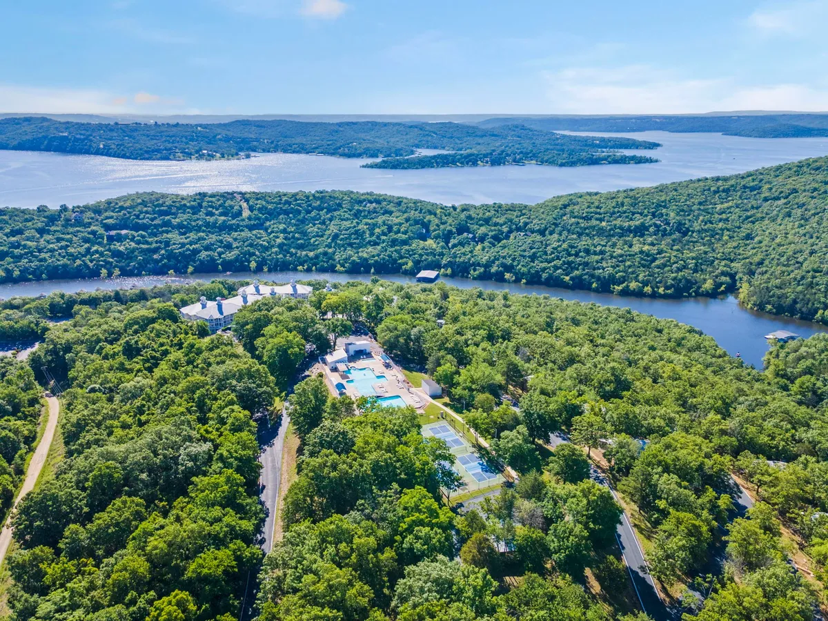 An expansive aerial view of the resort grounds featuring a swimming pool, tennis courts (which could also be used for pickleball or basketball), and lush landscaping leading to the lake’s edge. The scene highlights the resort’s comprehensive amenities and natural beauty.