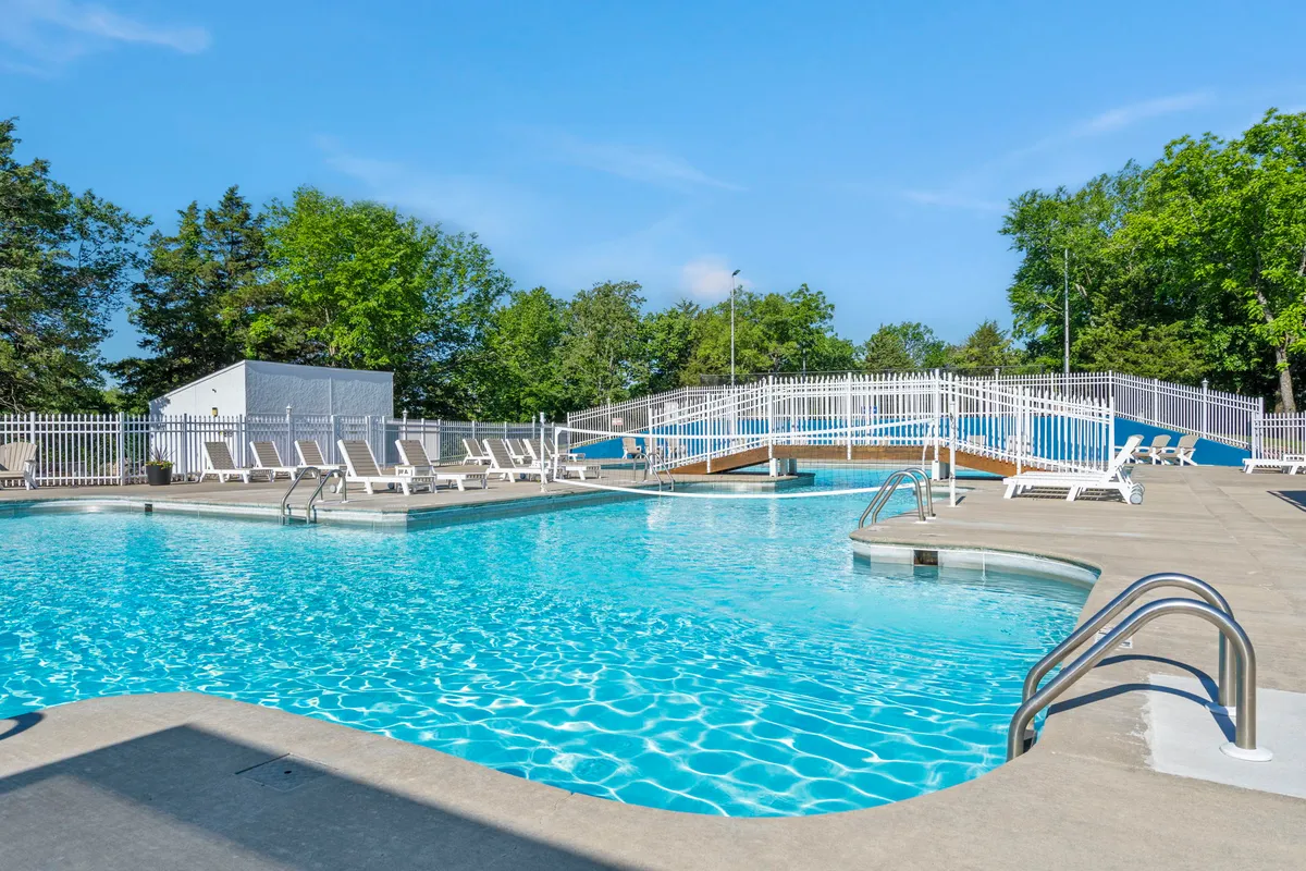A vibrant outdoor pool area with sparkling blue water, surrounded by lounge chairs, a charming bridge, and mature trees framing the space. The scene suggests a lively yet relaxed environment, ideal for swimming, sunbathing, or casual social gatherings.